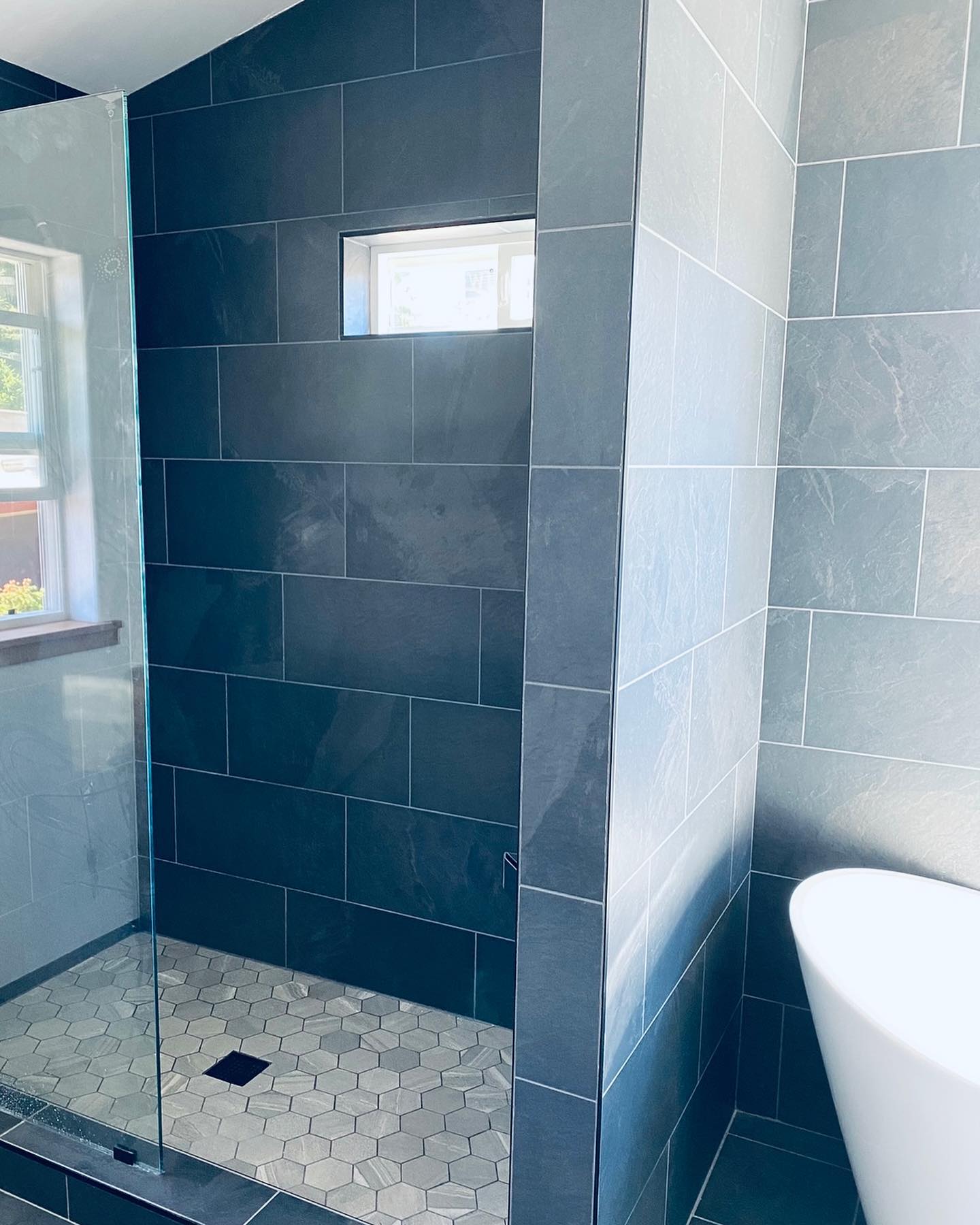 Modern custom bathroom featuring dark gray shower tiles, hexagonal floor tiles, glass shower door, and freestanding tub in Humboldt County.